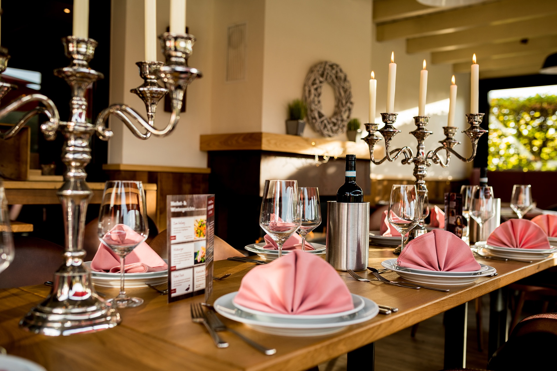 Elegante Tischdekoration in einem gehobenen Restaurant. Im Vordergrund stehen glänzende silberne Kerzenleuchter mit brennenden weißen Kerzen. Die Tische sind mit Weingläsern, gefalteten rosafarbenen Servietten auf weißen Tellern und Besteck sorgfältig gedeckt. Eine Flasche Wein und ein Weinbehälter stehen bereit. Im Hintergrund ist ein weiterer dekorativer Kranz an der Wand zu erkennen, der das stilvolle Ambiente unterstreicht.
