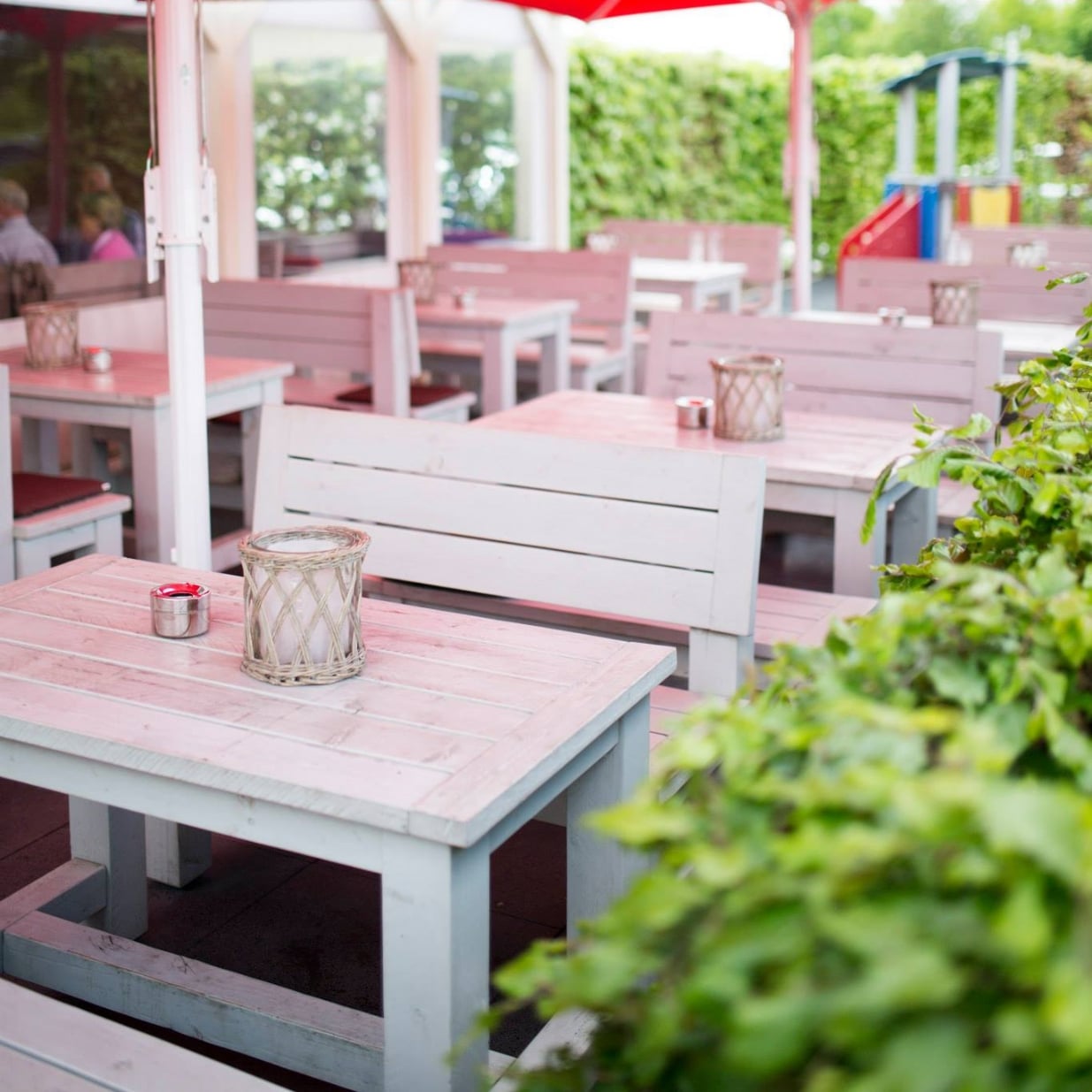 Leere Außenterrasse eines Restaurants mit stilvollen, pastellfarbenen Holztischen und Bänken, dekoriert mit einem windgeschützten Kerzenglas und einem kleinen Aschenbecher, umgeben von grüner Vegetation und einem Blick auf einen Spielplatz im Hintergrund.