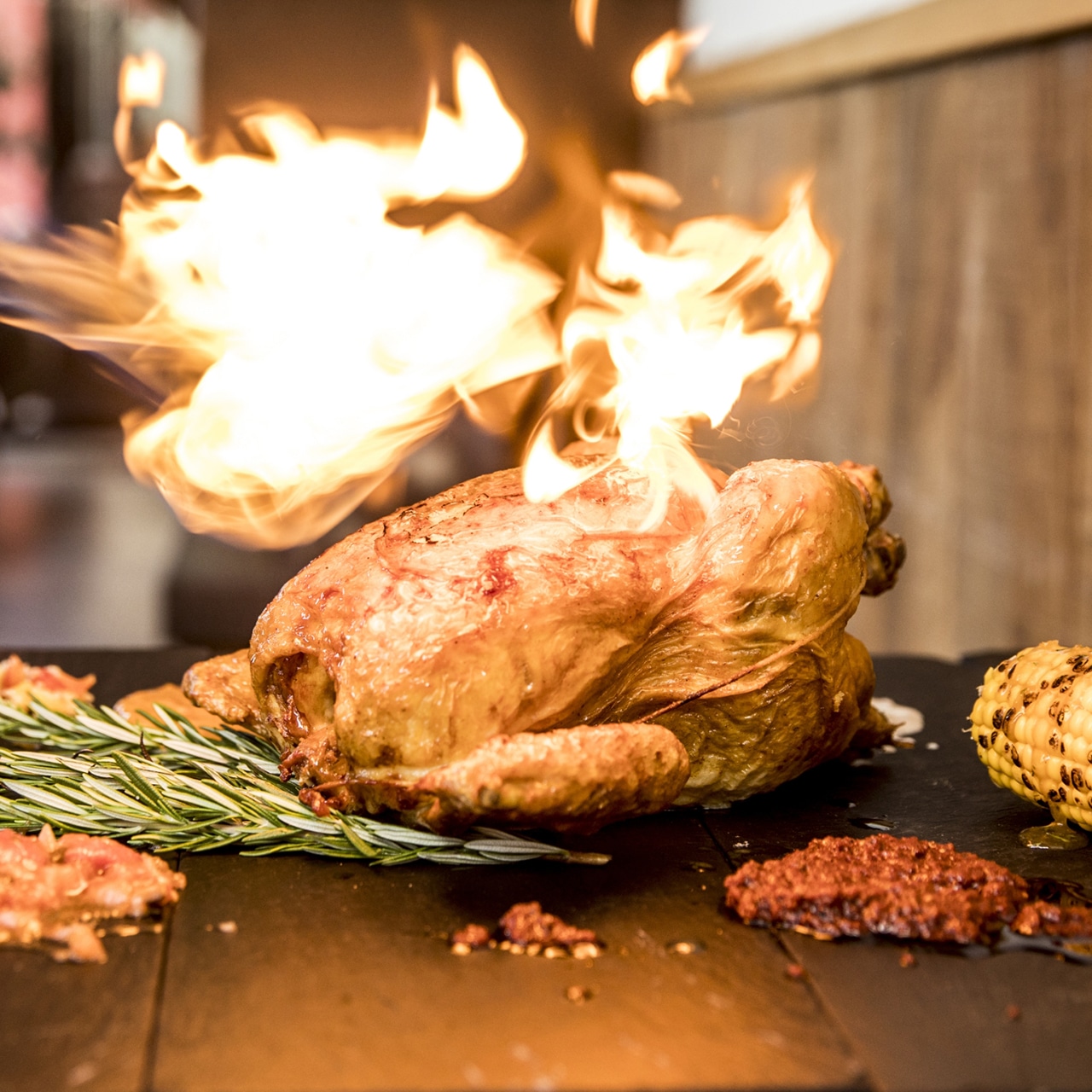 Spektakuläre Darstellung eines flambierten Hähnchens auf einem Holzbrett. Flammen umhüllen das goldbraun gebratene Hähnchen, das in der Mitte des Bildes platziert ist.