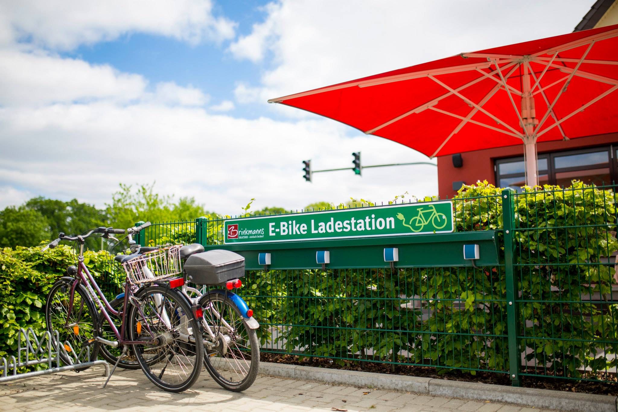Sonnenbeschienene E-Bike-Ladestation von 'Brinkmanns Braterei' neben geparkten Fahrrädern, mit einem großen roten Sonnenschirm im Hintergrund, der über einer grünen Hecke schwebt.