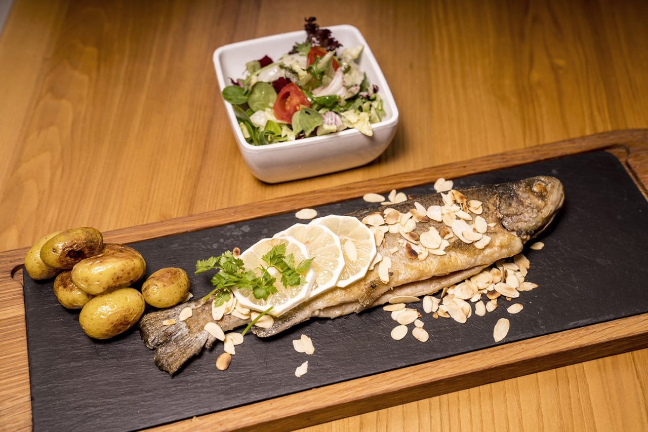 Ein appetitlich angerichteter Fisch, serviert auf einer Schiefertafel mit einer Seite von Bratkartoffeln und einer Zitronenscheiben-Garnitur, bestreut mit Mandelsplittern.
