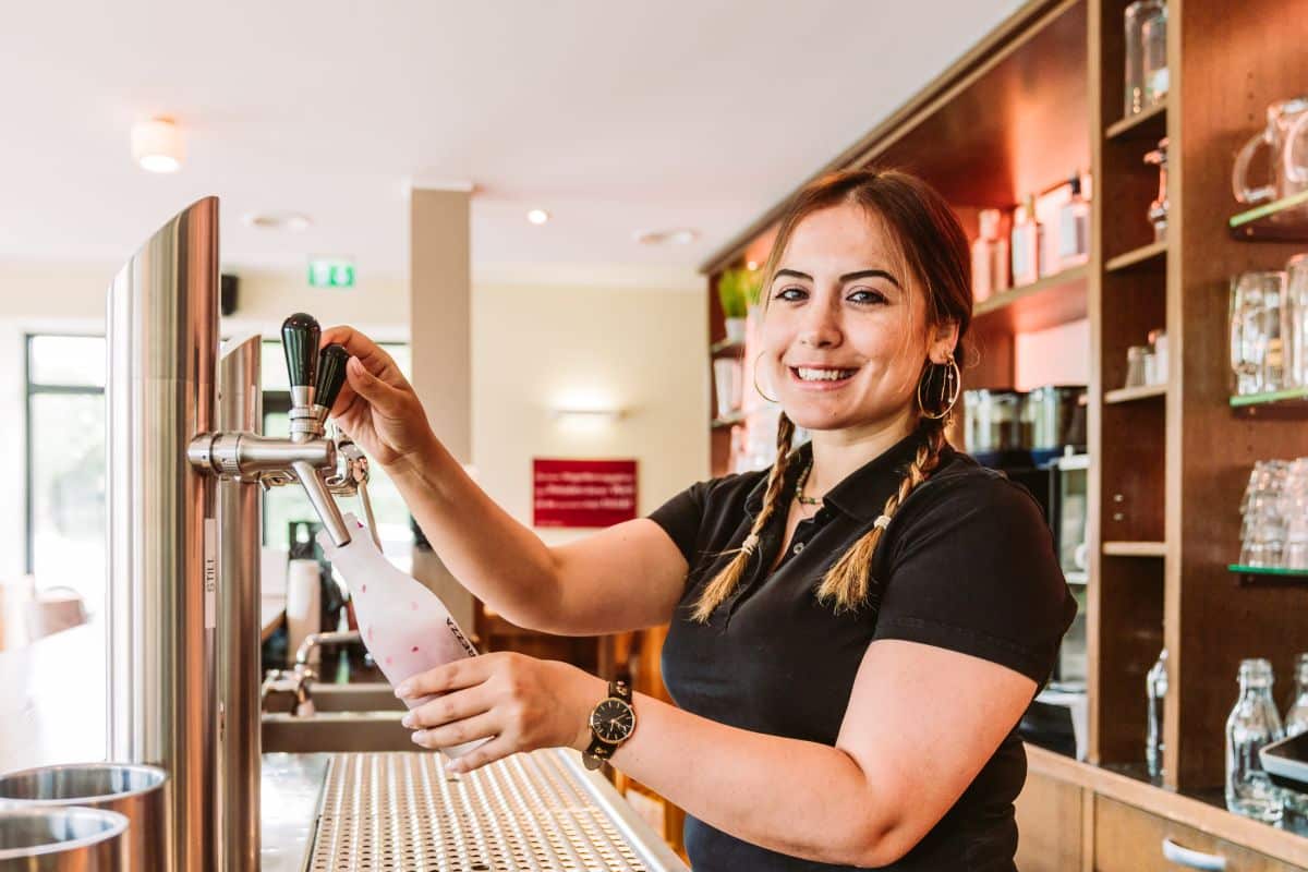 Fröhliche junge Kellnerin mit geflochtenen Haaren füllt ein Bierglas an einem Zapfhahn in einer Bar, mit einem Regal voller Gläser im Hintergrund.